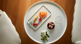 A white round table with a rectangular plate of assorted fruits, a small black plate with macarons, a glass of water, and a small bouquet of flowers at Avani Avani Palazzo Moscova Milan Hotel