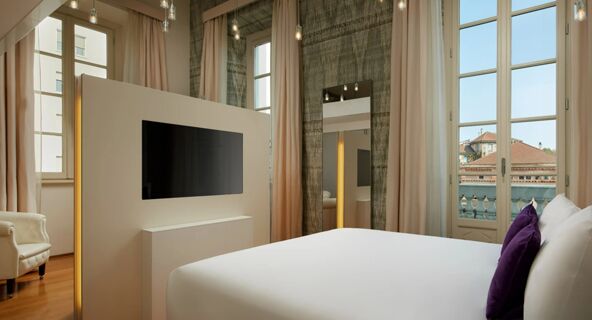 Bright hotel room at Avani Avani Palazzo Moscova Milan Hotel with a white bed, purple pillows, TV panel, large windows, and curtains
