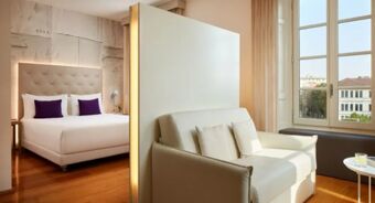 A bedroom with a large bed with purple pillows, a white sofa, wooden floor, and large windows at Avani Avani Palazzo Moscova Milan Hotel