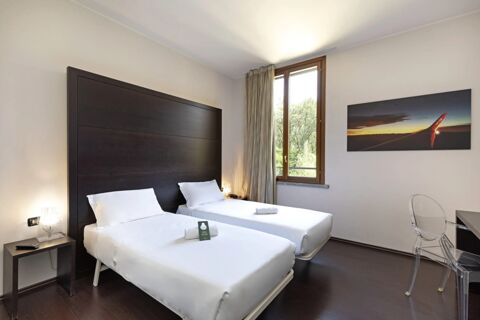 Two twin beds with white linens and towels, a dark wooden headboard, window with curtains, table, and chair in B&B Hotel Malpensa Lago Maggiore room