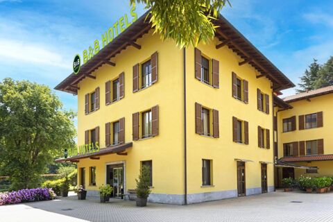 Yellow three-story building with brown shutters, green trees, flowers, and B&B Hotel Malpensa Lago Maggiore signage on top