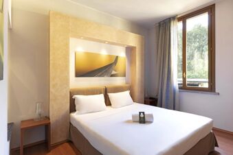Double bed with white linens and pillows under a framed abstract artwork in a B&B Hotel Malpensa Lago Maggiore room