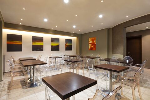 Dining area with dark wood square tables, transparent plastic chairs, ceiling spotlights, and sunset wall art at B&B Hotel Malpensa Lago Maggiore