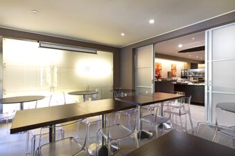 Interior dining area of B&B Hotel Malpensa Lago Maggiore with dark wooden tables, transparent chairs, and a buffet counter visible through partially open sliding doors
