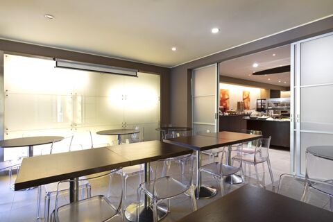 Interior dining area of B&B Hotel Malpensa Lago Maggiore with dark wooden tables, transparent chairs, and a buffet counter visible through partially open sliding doors