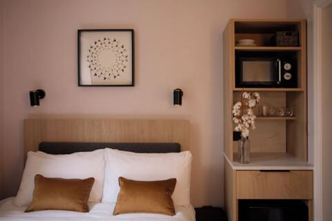 A double bed with white sheets and brown pillows under a framed geometric artwork and a wooden shelving unit with a microwave and vase at Bob W London Holland Park