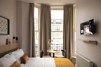 A hotel room at Bob W London Holland Park with a bed, white and brown pillows, a small round table with two chairs, and a wall-mounted TV