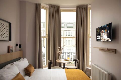 A hotel room at Bob W London Holland Park with a bed, white and brown pillows, a small round table with two chairs, and a wall-mounted TV