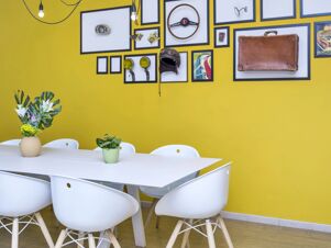 White chairs around a rectangular table with two small plant pots and a yellow wall with framed vintage car parts at B&B Hotel Milano-Monza