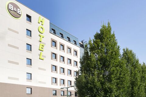 The exterior of B&B Hotel Milano-Monza featuring a beige and white facade with green hotel signage and a large tree on the right side