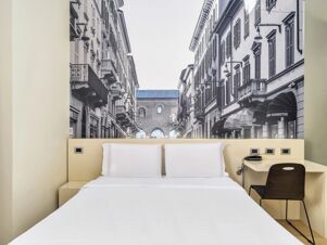 White bed with two pillows under a large black and white cityscape photograph and a small desk and chair at B&B Hotel Milano-Monza