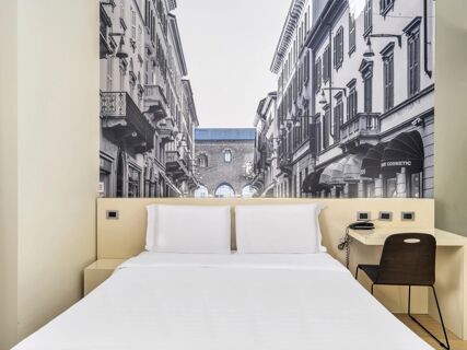 White bed with two pillows under a large black and white cityscape photograph and a small desk and chair at B&B Hotel Milano-Monza
