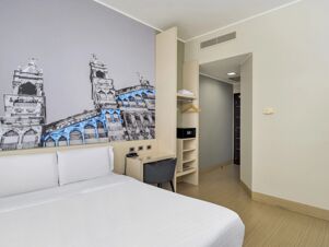 Hotel room at B&B Hotel Milano-Monza with a white bed, black and white wall art, desk, chair, and open closet with shelves and hangers