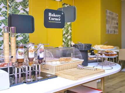 Buffet area at B&B Hotel Milano-Monza with jam dispensers, ice cream cones, bread, tarts, and hanging Bakery Corner signs