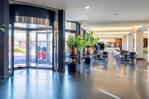 Lobby area of B&B Hotel Expo Verona Villafranca featuring a revolving glass door, potted plants, seating area, and a bar counter in the background