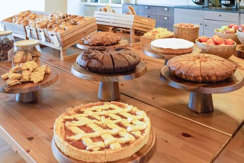 A wooden table displays various cakes, cookies, croissants, and bowls of apples and fruit at B&B Hotel Expo Verona Villafranca