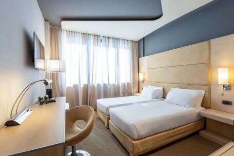 Two single beds with beige upholstery and white linens in a hotel room with desk, chair, TV, and window at B&B Hotel Expo Verona Villafranca