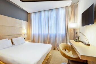 Hotel room at B&B Hotel Expo Verona Villafranca with a bed, beige headboard, bedside lamp, curtains, wall-mounted TV, and desk with chair