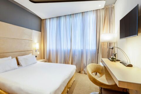 Hotel room at B&B Hotel Expo Verona Villafranca with a bed, beige headboard, bedside lamp, curtains, wall-mounted TV, and desk with chair