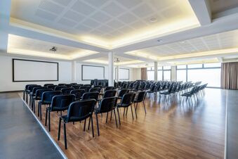 Conference room with multiple rows of black chairs, two large projector screens, and wood flooring at B&B Hotel Expo Verona Villafranca
