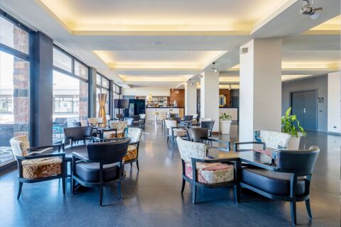 Spacious lobby area with patterned and black leather chairs, large windows, plants, and bar counter at B&B Hotel Expo Verona Villafranca