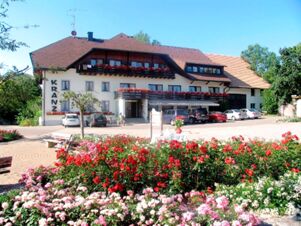 Gasthof Kranz is a multi-story white building with balconies decorated with red flowers and a parking area in front with several cars