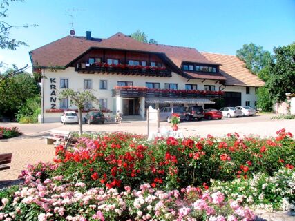 Gasthof Kranz is a multi-story white building with balconies decorated with red flowers and a parking area in front with several cars