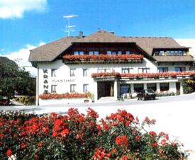 У отеля Gasthof Kranz традиционный белый фасад с деревянной крышей, множество цветочных балконов и красные цветы на переднем плане под солнечным небом.