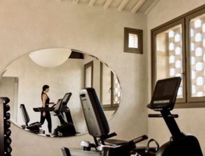 A woman using a treadmill reflected in an oval mirror inside the gym at COMO Castello Del Nero Hotel & Spa with exercise bikes nearby