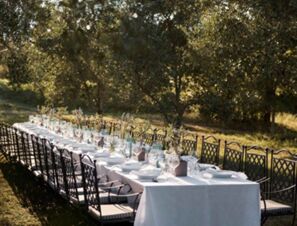 Long outdoor dining table with white tablecloth, glassware, and chairs under trees at COMO Castello Del Nero Hotel & Spa