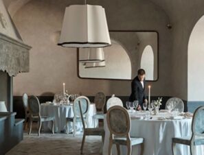Dining area with round tables covered in white tablecloths, a man setting a table, large pendant lights, and a mirror at COMO Castello Del Nero Hotel & Spa