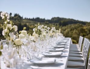 Long outdoor dining table set with white plates, glassware, and flower centerpieces at COMO Castello Del Nero Hotel & Spa surrounded by greenery