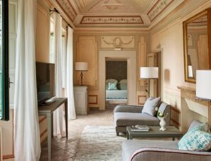 Living area with grey sofas, coffee table, large windows with curtains, and ornate walls at COMO Castello Del Nero Hotel & Spa
