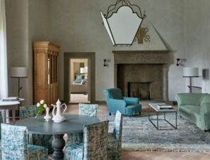 Living area with teal upholstered chairs, blue patterned dining chairs around a table, and a large stone fireplace at COMO Castello Del Nero Hotel & Spa