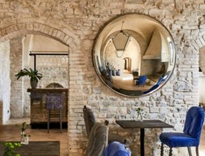 Dining area with stone walls, wooden tables, blue and gray chairs, a large round mirror, and a window at COMO Castello Del Nero Hotel & Spa
