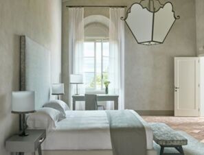 Bedroom in COMO Castello Del Nero Hotel & Spa featuring a large bed, bedside tables with lamps, a window with sheer curtains, and a bench