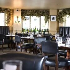 Interior dining area at Hotel du Vin Cheltenham with dark leather chairs, wooden tables, and wine bottles displayed near windows