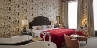 Hotel du Vin Cheltenham bedroom with double bed covered by a red blanket, floral wallpaper, two bathtubs, and a rocking horse near large windows
