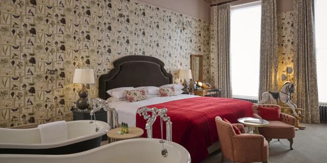 Hotel du Vin Cheltenham bedroom with double bed covered by a red blanket, floral wallpaper, two bathtubs, and a rocking horse near large windows