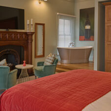 Bedroom interior at Hotel du Vin Cheltenham featuring a red bedspread, fireplace, two green chairs, and a freestanding bathtub near a window with blinds