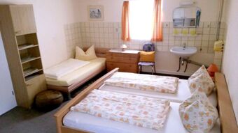 Ein einfach eingerichtetes G&auml;stezimmer mit zwei Einzelbetten, einem weiteren Bett, Waschbecken und M&ouml;beln in der Pension Arnold.