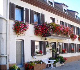 Fassade der Pension Arnold mit zahlreichen bunten Blumen an Fenstern und Balkon, gem&uuml;tlicher Tisch mit Stuhl davor.
