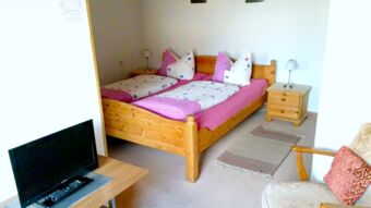 Schlicht eingerichtetes Zimmer der Pension Arnold mit Holzbett, Nachtisch, Teppichen und Fernseher auf einem kleinen Tisch.