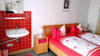 Helles Zimmer in der Pension Arnold mit zwei Betten, roten Bettw&auml;sche und einem Waschbecken an der Wand mit roten Fliesen.