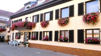 Au&szlig;enansicht der Pension Arnold mit gelb gestrichener Fassade, schwarzen Fensterl&auml;den und zahlreichen bunten Blumenarrangements an Fenstern und Balkon.