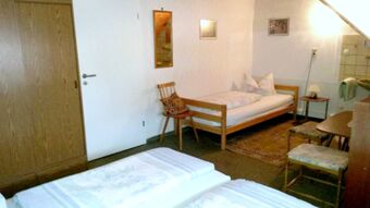 Gem&uuml;tliches Zimmer mit Einzelbett, Stuhl, Teppich und Tisch in der Pension Arnold mit schlichtem Holzdesign und heller Wand.