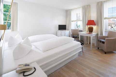 Bright hotel room at Landhotel Krone with double bed, white linens, two armchairs, table with red lamp, TV, and windows with curtains