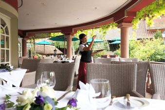 A waitress holding a glass and tray stands on the outdoor terrace of Landhotel Krone with wicker chairs and set tables visible