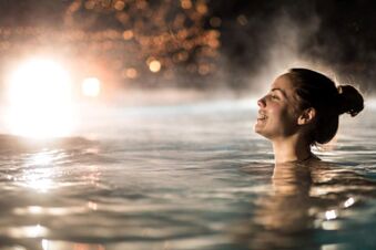 A woman with a bun is smiling and relaxing in a steamy pool at night with glowing lights near Landhotel Krone