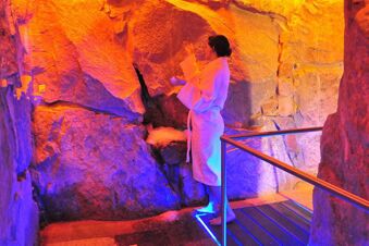 A woman in a white bathrobe stands barefoot on a metal platform inside a colorful illuminated stone cave at Landhotel Krone